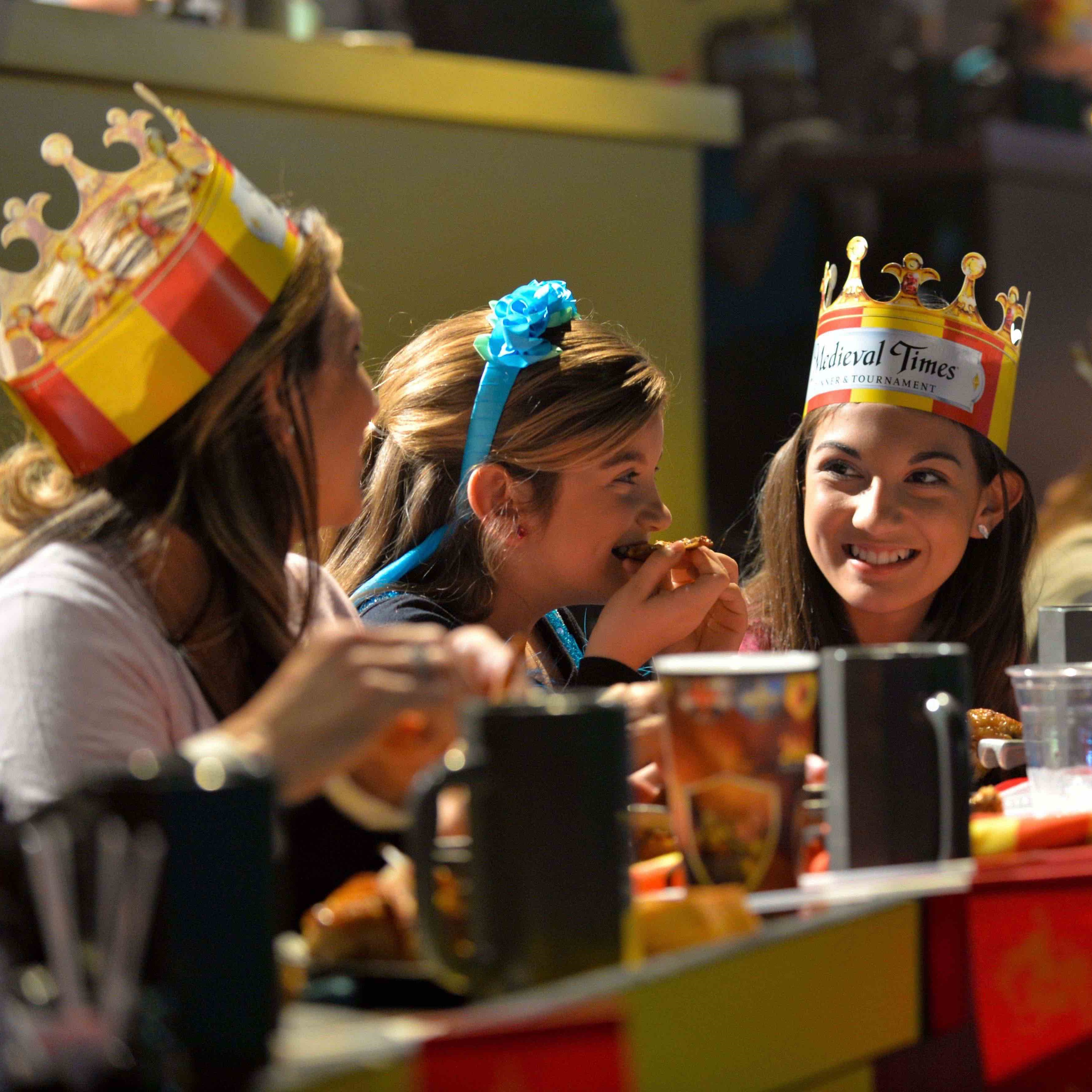 Three young girls smiling at each other in the arena tables wearing crowns 